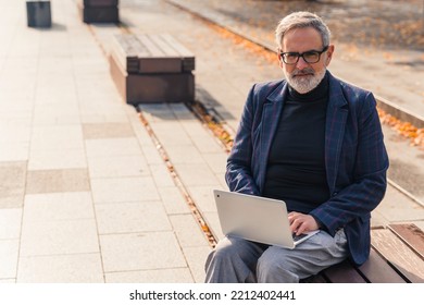 Remote Working Concept. Adult Caucasian Gray-haired Bearded Businessman In Formal Clothes Benefiting By Beautiful Sunny Autumn Weather And Working Outside With His Laptop. Looking At Camera. High