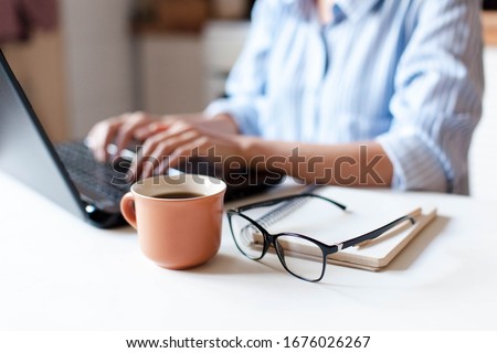 Remote working from home. Freelancer workplace in kitchen with laptop, cup of coffee, spectacles. Concept of distance learning, isolation, female business, shopping online. Close up of woman hands.