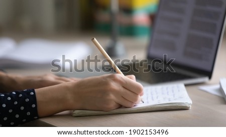 Remote working or learning. Close up of young woman hands making notes in paper copybook on desk by computer screen. Female student or pupil writing out information studying lecture lesson from home