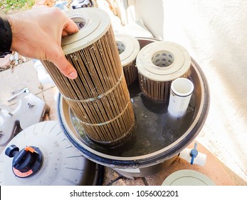 Removing A Dirty Reusable Washable Pleated Reinforced Polyester Cartridge Pool Filter From Its Murky Water-filled Housing, Pending Maintenance Cleaning.  Other Filters, Tools, Valves Are Visible.