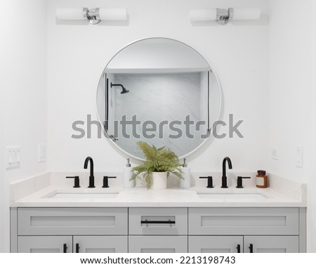 A renovated bathroom with a grey vanity cabinet, circular mirror with a view to a shower, and back faucets.