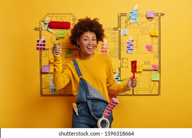 Renovation At Home. Positive African American Woman Holds Paint Brush And Roller, Busy With Painting, Thinks About House Improvement, Dressed In Jean Overalls, Creative Sketch On Yellow Wall