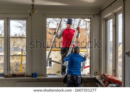 A renovation project in progress as two workers replace an old window. One man passes the frame from the outside while the other prepares the opening from inside the home.