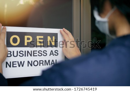 Reopening for business adapt to new normal in the novel Coronavirus COVID-19 pandemic. Rear view of business owner wearing medical mask placing open sign “OPEN BUSINESS AS NEW NORMAL” on front door.