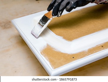 Repainting Kitchen Cabinet Doors With White Chalk Paint Indoors At Home. Giving Old Kitchen New Look Concept. Hand Holding A Paint Brush Tool With Paint Against Old Cupboard Door.