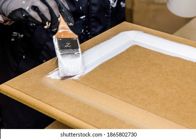 Repainting Kitchen Cabinet Doors With White Chalk Paint Indoors At Home. Giving Old Kitchen New Look Concept. Hand Holding A Paint Brush Tool With Paint Against Old Cupboard Door.