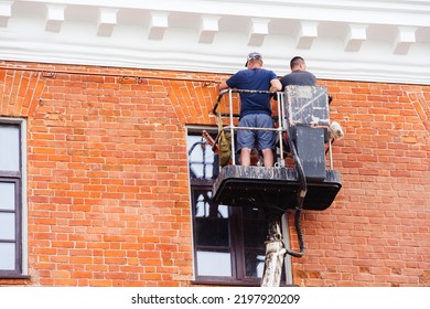 Repair Of The Facade Of A Brick House. Two Workers On An Aerial Platform Inspect The Wall And Identify Defects After Major Repairs. Unrecognizable Person