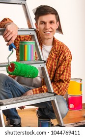 Repair Home Man Holding Paint Roller For Wallpaper. Tired And Frustrated Male In Newspaper Cap Renovation Apartment. Paint Bank And Roll Paper Background On Isolated. Stepladder In Foreground.