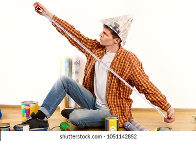 Repair Home Man Holding Paint Roller For Wallpaper. Tired And Frustrated Male In Newspaper Cap Renovation Apartment. Person Measures Room By Centimeter. Young Student Builder.