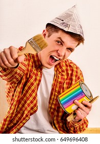 Repair Home Man Holding Paint Roller For Wallpaper. Tired And Frustrated Mad Male In Newspaper Cap Renovation Apartment. Paint Bank And Paint Brush In Hand . Worker-builder Student.