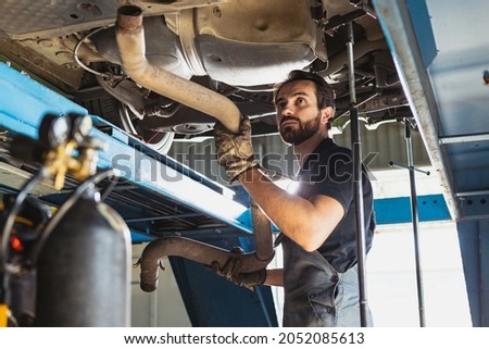 Repair of machinery. Portrait of young bearded man, male auto mechanic in dungarees working at car service station, indoors. Concept of labor, business, caree, job. Nonprofessional occupations