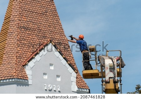 Repair and reconstruction of the old roof made of red tiles of a century-old building. The worker is in the basket of a crane and works at a height in dangerous conditions. Roof repair on a sunny day