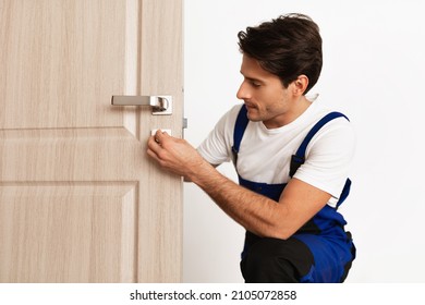 Repair Service Concept. Portrait of smiling handsome young locksmith in blue uniform installing door knob on front wooden entrance door at home or office workplace, isolated on white studio background