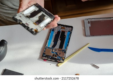 Repair Of Smartphones Concept. A Man Holding A New Display In His Hand. A Disassembled Phone With An Old, Broken Display Lying On The Table. Part Of A Series. Indoors. Selective Focus.