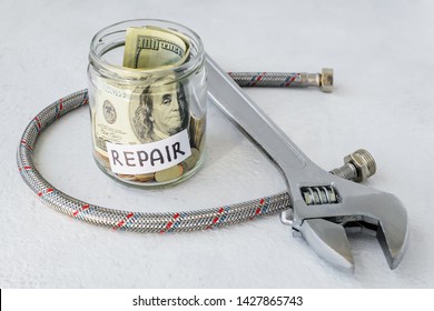 Repair word, coins and dollar bill in glass jar near flexible metal braided hose for plumbing taps and metal adjustable wrench on a white background. Saving money for repair concept. 
