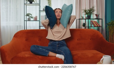 Repair work at neighbours concept. Irritated woman sit on couch cover ears with pillows annoyed by noisy neighbors suffer from headache wishes silence. Thin walls at home flat without sound insulation