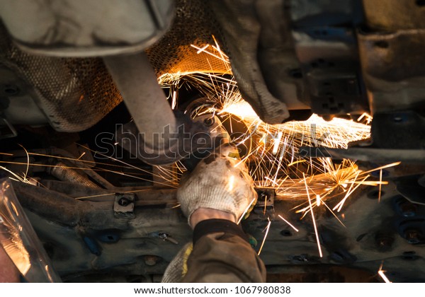 repairing of corrugation muffler of exhaust\
system in car workshop - serviceman cleans the muffler pipe on car\
by angle grinder