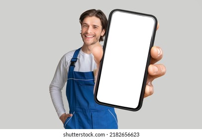 Repairing Service App. Young Handyman In Blue Workwear Showing Modern Mobile Phone With White Blank Screen And Smiling, Posing Over Grey Background With Copy Space, Collage, Mockup