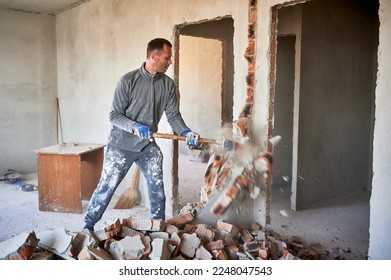 Repairman crashing brick wall with sledgehammer, pieces of brick flying away from strong blow. Destruction of partitions in flat during overhaul.