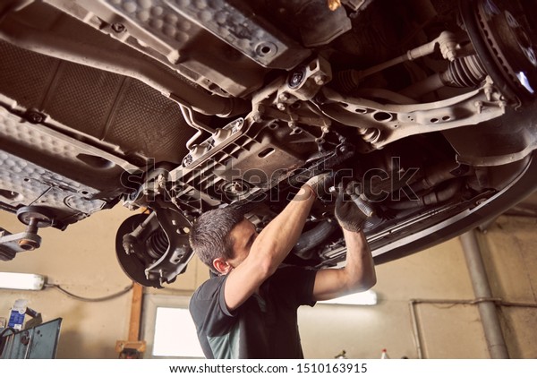 Repairman fixing car in garage. Experienced\
specialist car mechanic standing under lifted car during repair and\
maintenance process in repair station. Workshop for automobile\
checking up and\
repairing
