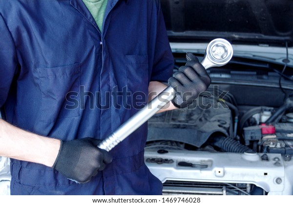 Repairman is repairing car at service station. Closeup\
mechanic hands in gloves are holding big steel wrench. Vehicle with\
open hood on background.  Modern auto repair shop with equipments\
and tools. 