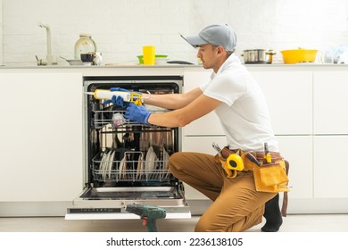 Repairman in uniform repairs dishwasher in kitchen. Young man specialist unscrews parts with screwdriver checking state of trays for dish side view.