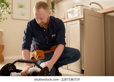 Repairman in uniform taking detail from his bag to repair sink in domestic kitchen