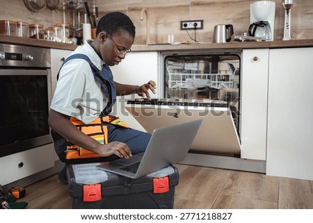 Repairman using laptop while fixing dishwasher in kitchen.
