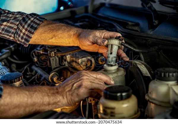 Repairman\'s male\
hands checking level of the brake fluid. Vehicle fitter inspecting\
used car engine. Car components, belts, hoses, labor arm close up\
in the open hood of the car.\
