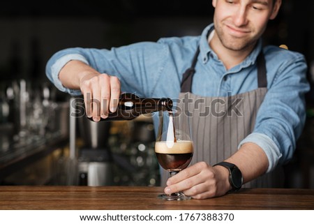 Repeat the order. Bartender in apron and denim shirt, pours dark beer in glass on wooden bar counter in interior of pub