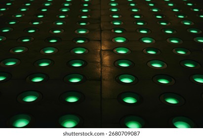 A repeating pattern of circular indentations, each emitting a soft green light, creates a mesmerizing visual on a dark, textured surface. The perspective emphasizes the depth and repetition.