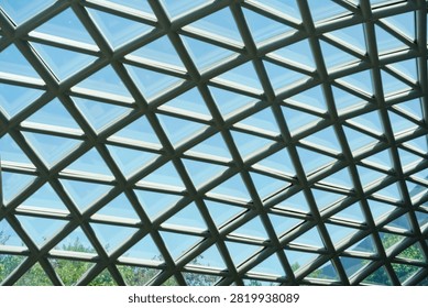 A repeating pattern of triangular glass panels and metal framework creates a modern architectural ceiling against a bright blue sky