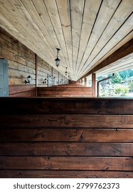 A repeating single-point perspective view of a long wooden balcony or veranda corridor of a rustic hotel or resort. The scene is constructed of natural wood. The foreground focuses on a dark brown horizontal wooden partition. Similar partitions are repeated in a pattern along the long terrace, dividing individual guest rooms. The left wall consists of horizontal log-style siding with a closed gray wooden door. The ceiling is made of light pine planks that converge sharply to a central point of elevation. A neat row of vintage-style black pendant lights is installed along the ceiling, echoed by matching wall lights near each door. The open right side of the veranda offers a view of the bright, sunny green garden, courtyard and trees beyond. Soft, natural daylight fills the hallway, highlighting the rich textures, grains and knots of the wood.