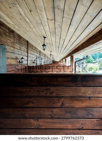 A repeating single-point perspective view of a long wooden balcony or veranda corridor of a rustic hotel or resort. The scene is constructed of natural wood. The foreground focuses on a dark brown horizontal wooden partition. Similar partitions are repeated in a pattern along the long terrace, dividing individual guest rooms. The left wall consists of horizontal log-style siding with a closed gray wooden door. The ceiling is made of light pine planks that converge sharply to a central point of elevation. A neat row of vintage-style black pendant lights is installed along the ceiling, echoed by matching wall lights near each door. The open right side of the veranda offers a view of the bright, sunny green garden, courtyard and trees beyond. Soft, natural daylight fills the hallway, highlighting the rich textures, grains and knots of the wood.