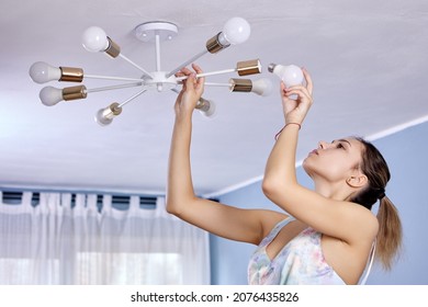 Replacing Electric Light Bulb In Ceiling Lighting Fixture. Slender European Woman 24 Years Old Changes Lamp In Chandelier.