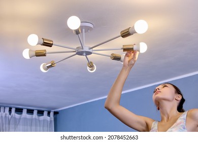 Replacing An LED Light Bulb In Ceiling Chandelier At Home, Young White Woman Holding New Light Lamp In Her Hand.