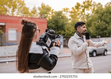 Reporter holding a microphone and gesturing while presenting news outdoors, with a camerawoman filming the broadcast for media coverage and journalism