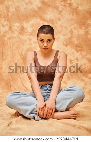 representation of body, casual wear, curvy, young and tattooed woman in jeans and crop top sitting on mottled beige background, personal style, self-acceptance, generation z, denim fashion, tattooed