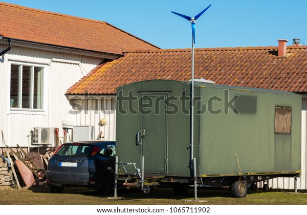 Repurposed military trailer outside a house.\
Small wind power generator supply electricity. Car parked beside\
trailer. Logo and registration\
removed.