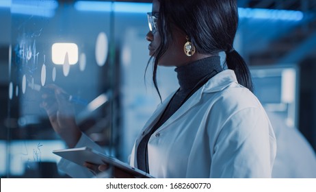 In The Research Laboratory Smart And Beautiful African American Female Scientist Wearing White Coat And Protective Glasses Writes Formula On Glass Whiteboard, References Her Tablet Computer