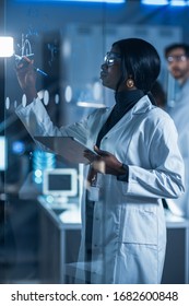 In The Research Laboratory Smart And Beautiful African American Female Scientist Wearing White Coat And Protective Glasses Writes Formula On Glass Whiteboard, References Her Tablet Computer