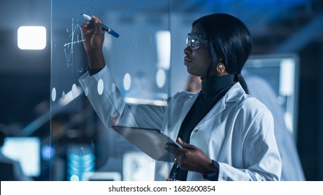 In the Research Laboratory Smart and Beautiful African American Female Scientist Wearing White Coat and Protective Glasses Writes Formula on Glass Whiteboard, References Her Tablet Computer