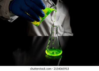 A Research Scientist Experimenting With A Green Fluorescent Droplets In A Glass Conical Flask In Dark Biomedical Laboratory For Health Care Medicine Development. Copy Space Black Background