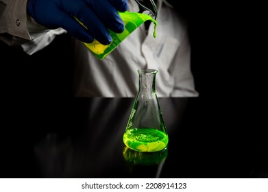 A Research Scientist Experimenting With A Green Fluorescent Droplets In A Glass Conical Flask In Dark Biomedical Laboratory For Health Care Medicine Development. Copy Space Black Background