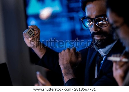 A researcher presents a microprocessor to colleagues, holding the small silicon chip up between his thumb and finger in a dark technology lab as the team leans in to study the hardware.