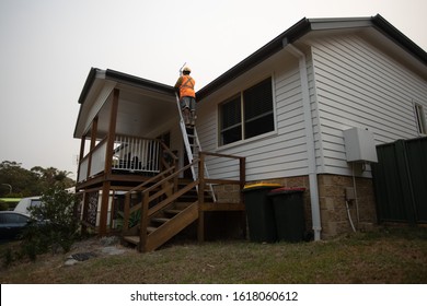 Resident From Sydney South Coast Wearing Helmet Climbing Standing On Industrial Safety Ladder While Cleaning House Gatter During Bushfire Season Sydney, Australia     