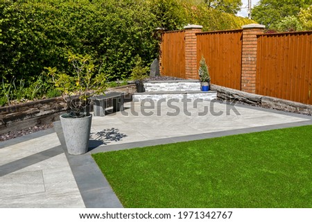 Residential back garden landscaped with light and dark grey porcelain paving slabs and artificial grass. No people.