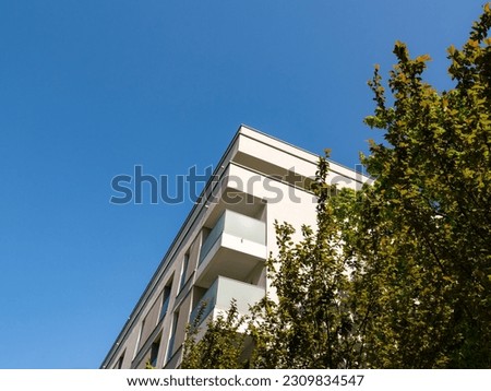 Residential building facade with balconies and glas railings. The sky is blue and clear. A tree with lush green leaves is in the front. The architecture is functional and sparse.