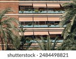Residential building in Valencia with vibrant orange-striped awnings and lush balcony plants. Palm trees frame the scene, adding a tropical touch to the urban landscape.