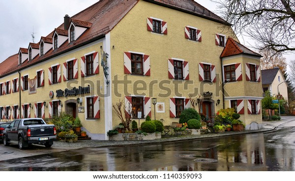 Residential house of a yellow color with white-red\
window shutters and red tile roof. Restaurant. Cars in a parking\
lot. Urban landscape. German Architecture. Germany, Zorneding –\
November 19, 2017 